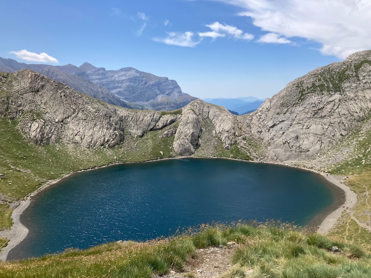 A serene mountain lake is depicted surrounded by rugged rocky terrain. The water appears calm, reflecting the sky's blue hues. Grass and small plants frame the foreground, leading up to the rocky slopes and distant mountain peaks in the background.