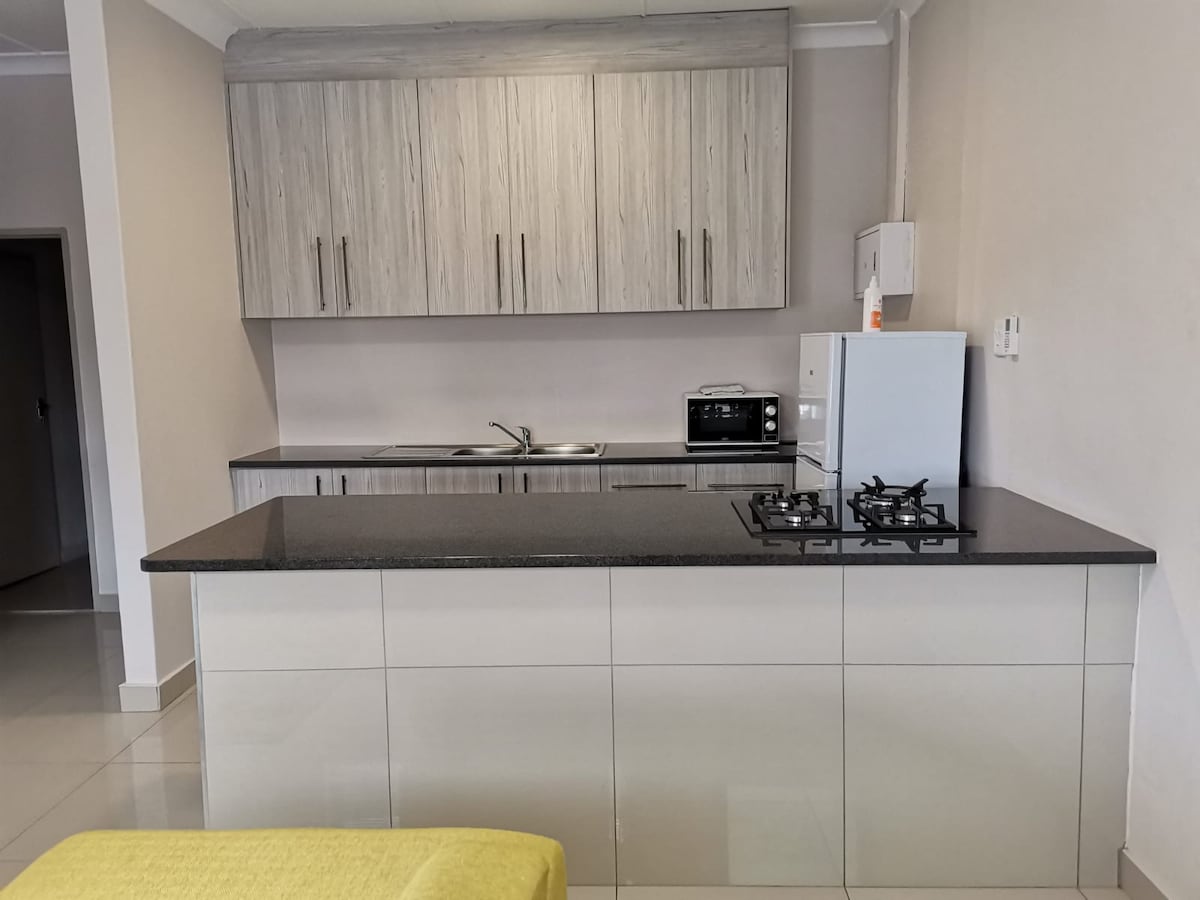A modern kitchen area features sleek cabinetry in light wood tones, complemented by a dark countertop. A gas stove and microwave are present, alongside a sink. The space is well-lit, with glossy tiles enhancing the contemporary ambiance.