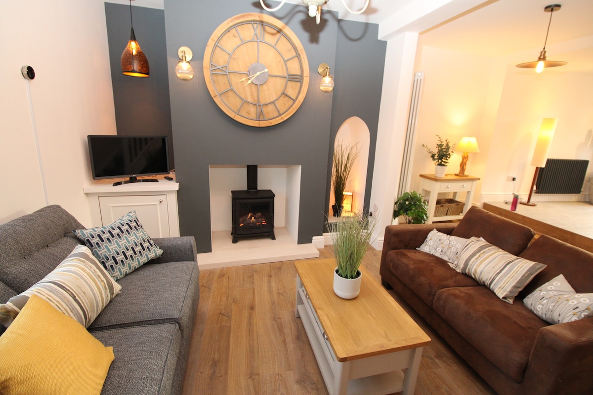 A cozy living area showcases two comfortable sofas, one in grey and one in brown, arranged around a wooden coffee table. A stylish clock and decorative lighting are featured on the wall, while a cast iron stove fireplace adds warmth to the space.