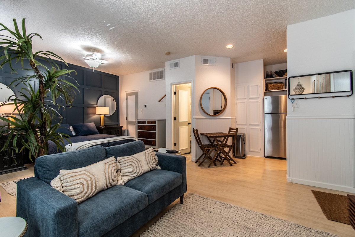 A stylish studio space features a cozy seating area with a blue sofa. The spacious bed is flanked by wall-mounted mirrors and complemented by warm lighting. A dining table with folding chairs is positioned near the kitchenette, enhancing the open and approachable layout of the room.