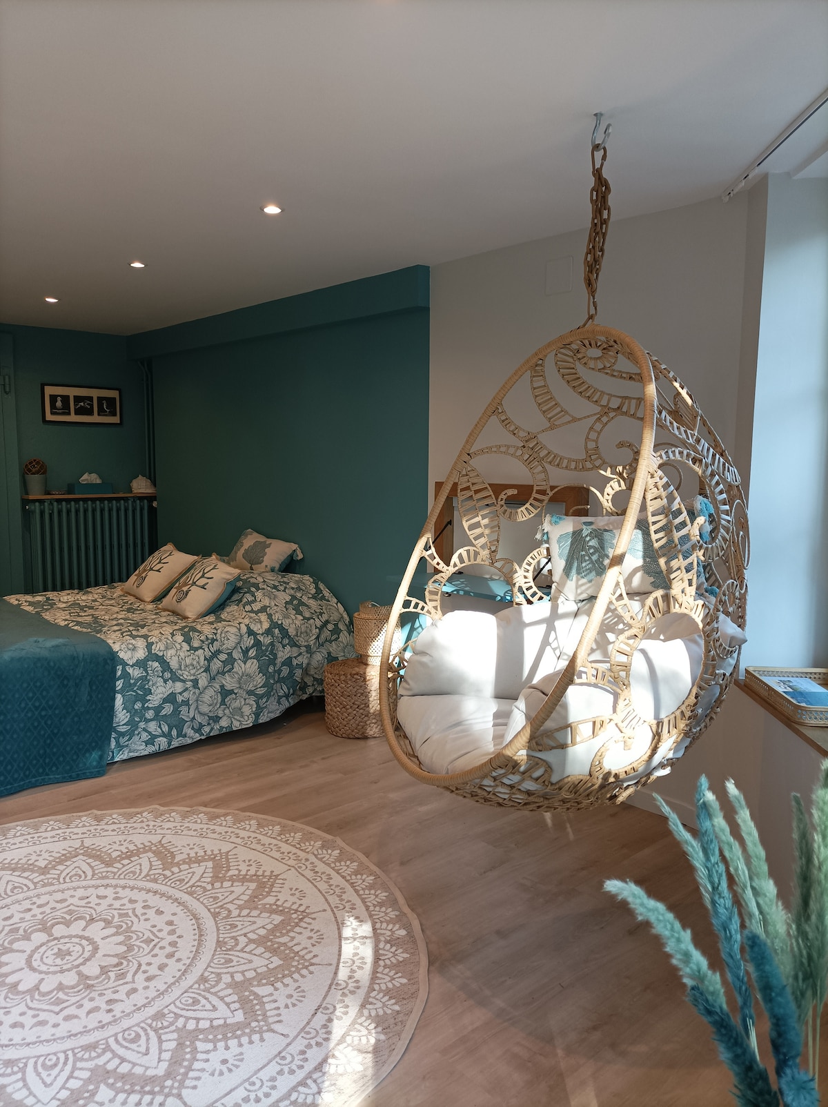 A hanging chair with intricate designs is featured in the room, complementing the serene blue walls. A spacious bed with patterned bedding occupies one side, while a round area rug adds texture to the wooden floor. Soft lighting enhances the calming atmosphere.