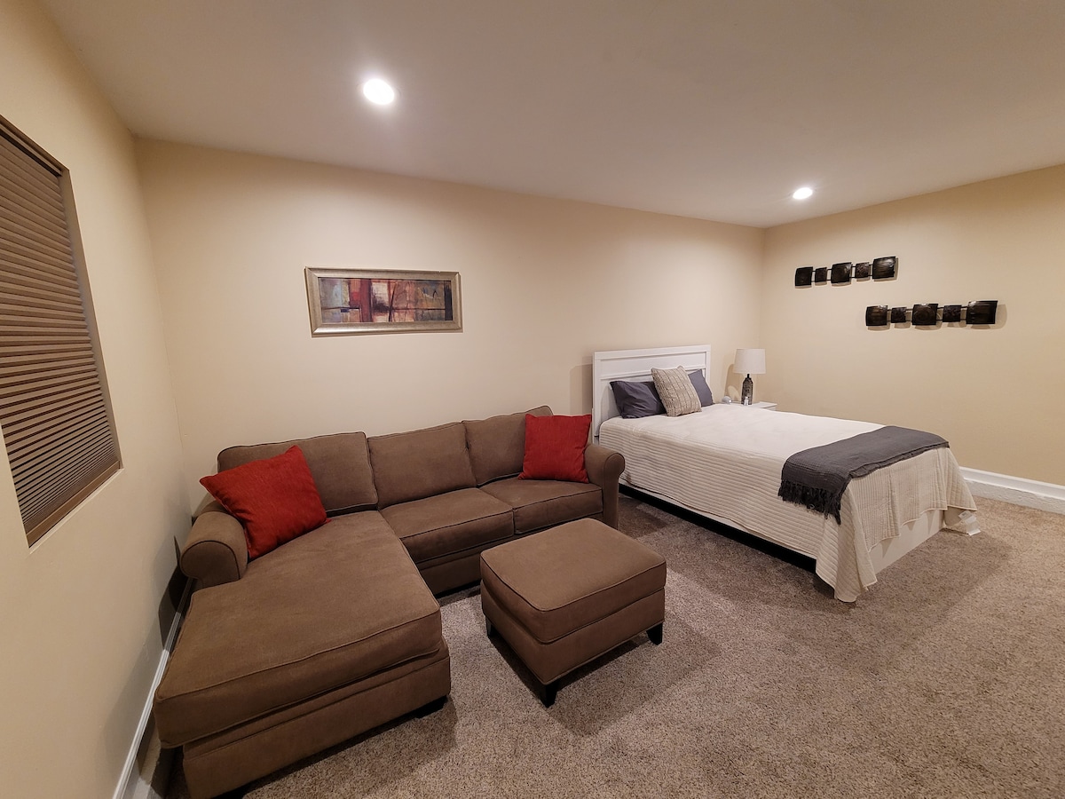 A spacious room is furnished with a plush brown sectional sofa adorned with red accent pillows and an ottoman. A large bed with neutral bedding is positioned nearby, and framed artwork adds subtle elegance to the wall, complemented by soft ceiling lights that enhance the inviting space.