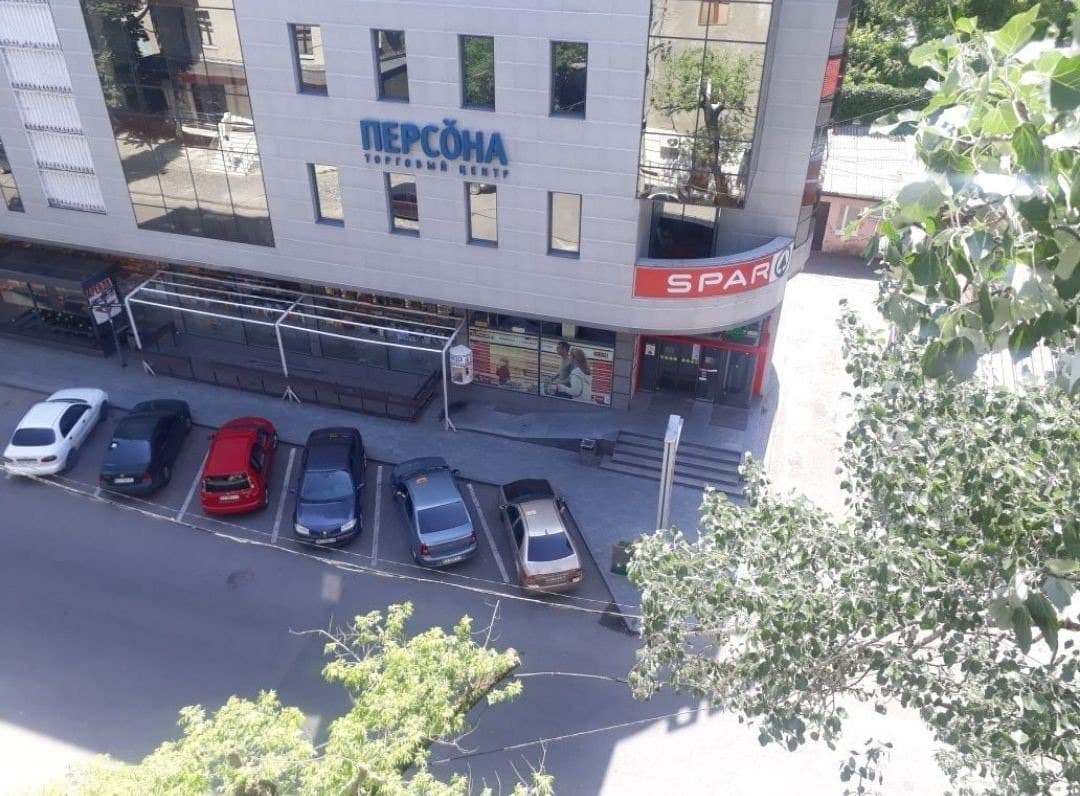 The image displays a street view featuring a modern building facade with large windows. The ground level shows retail signs, including 'PERSOÑA' and 'SPAR.' The foreground includes several parked cars along the roadside, with greenery visible around the scene.