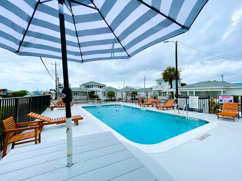 An outdoor pool area is highlighted by wooden lounge chairs arranged around the water. A large, striped umbrella provides shade. The surrounding fence and palm trees create a welcoming environment, with nearby cottages visible in the background.