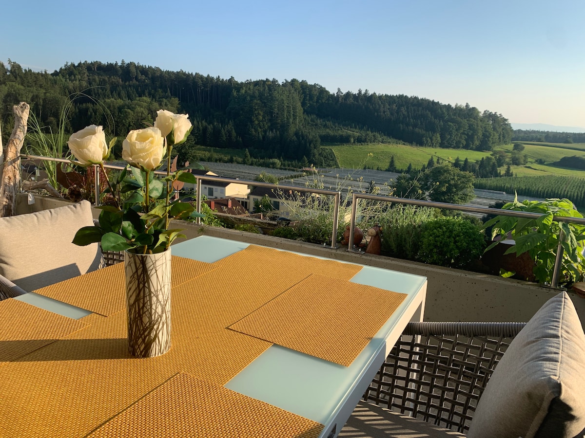 A dining table set on a terrace is prepared for meals, with a vase of fresh white roses at its center. A soft sunlight bathes the area, illuminating the surrounding greenery and distant hills, enhancing the serene atmosphere. Comfortable seating provides an inviting setting for relaxation.