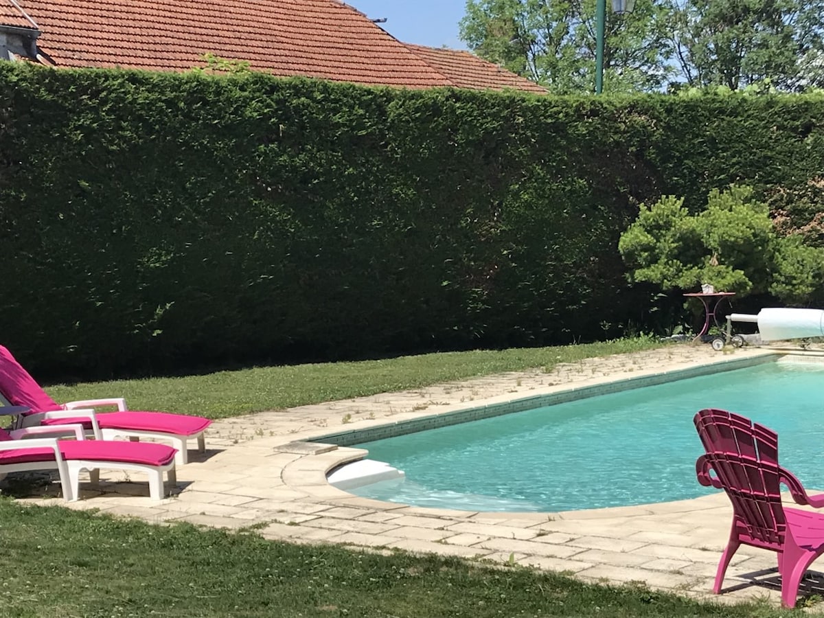 A serene outdoor area features a turquoise pool surrounded by a stone deck. Pink lounge chairs are positioned along the poolside, and a neatly trimmed hedge offers privacy. A small table is located nearby, enhancing the tranquil setting.