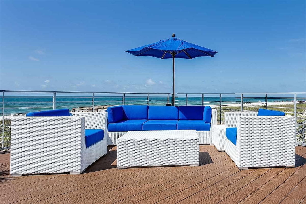 A patio area is furnished with a white wicker seating set featuring blue cushions. A table is positioned at the center. A large blue umbrella provides shade, and the view of the ocean and shoreline is clearly visible in the background.