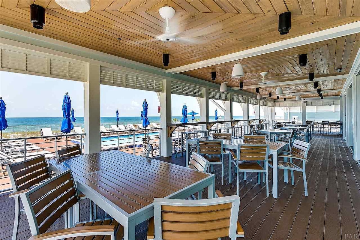 An outdoor dining area is shown, featuring several wooden tables and chairs arranged under a shaded roof. Blue umbrellas provide shade while offering a view of the ocean in the background, visible through large windows and open spaces.