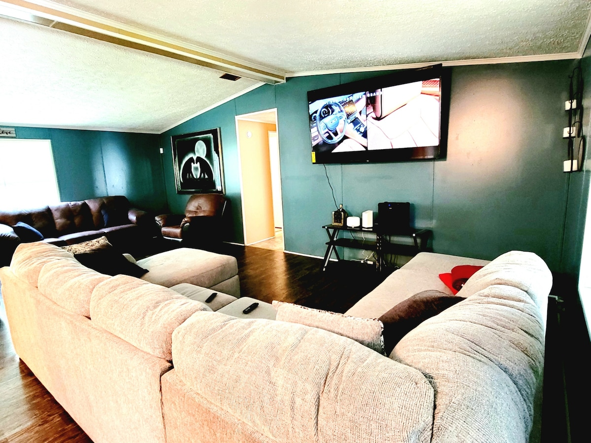 A spacious living area features a large sectional sofa in light grey, providing ample seating. A wall-mounted television is visible opposite the seating arrangement. A dark accent chair and a small table contribute to the cozy atmosphere, with teal walls enhancing the overall design.