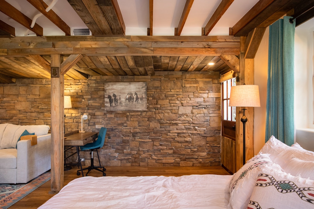 A spacious suite is highlighted by natural wood beams and a stone accent wall. Soft lighting is provided by a floor lamp next to a cozy couch. A desk with a chair is positioned nearby, and large windows allow for ample natural light.