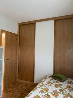 A well-organized bedroom features light wooden paneling and a spacious bed with a floral patterned duvet. A green cushion rests on the bed, while a closet occupies part of the wall, and a doorway leads to another area of the space.