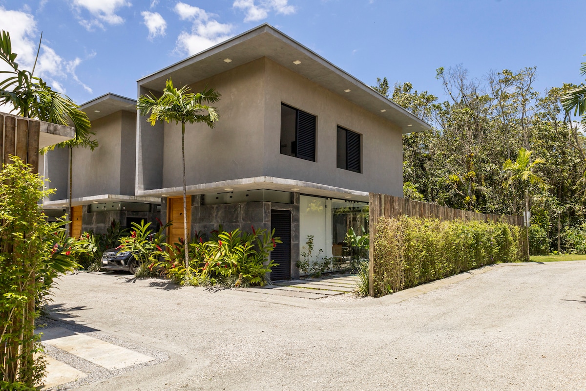 A modern two-story building is framed by lush greenery and tropical plants. The property features large windows and a spacious driveway leading to a private garage, providing ample parking space. Natural lighting enhances the architectural details of the exterior.