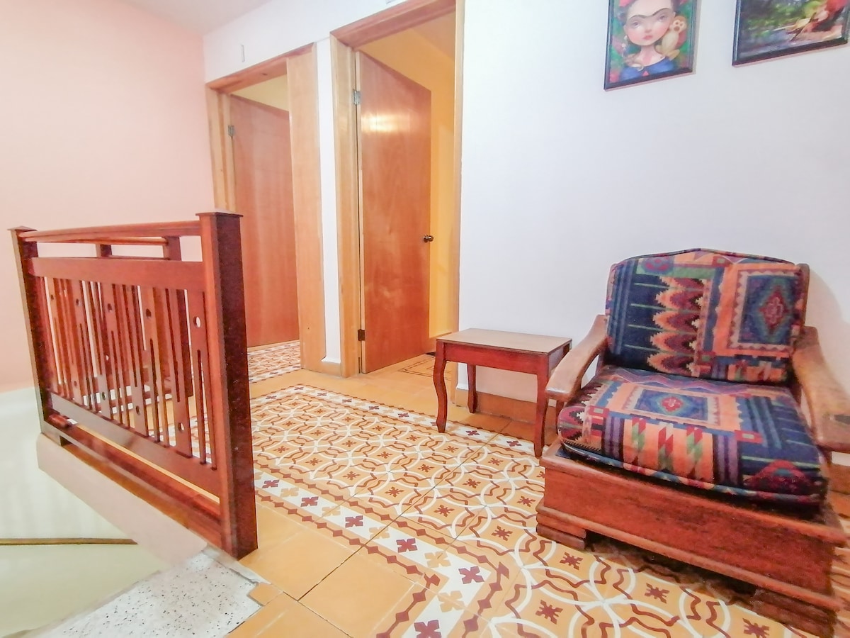 A cozy hallway features a wooden railing and a patterned floor. A comfortable chair with vibrant upholstery is positioned beside a small table. Two doorways lead to private rooms, while framed artwork adorns the wall, adding a touch of personality to the space.