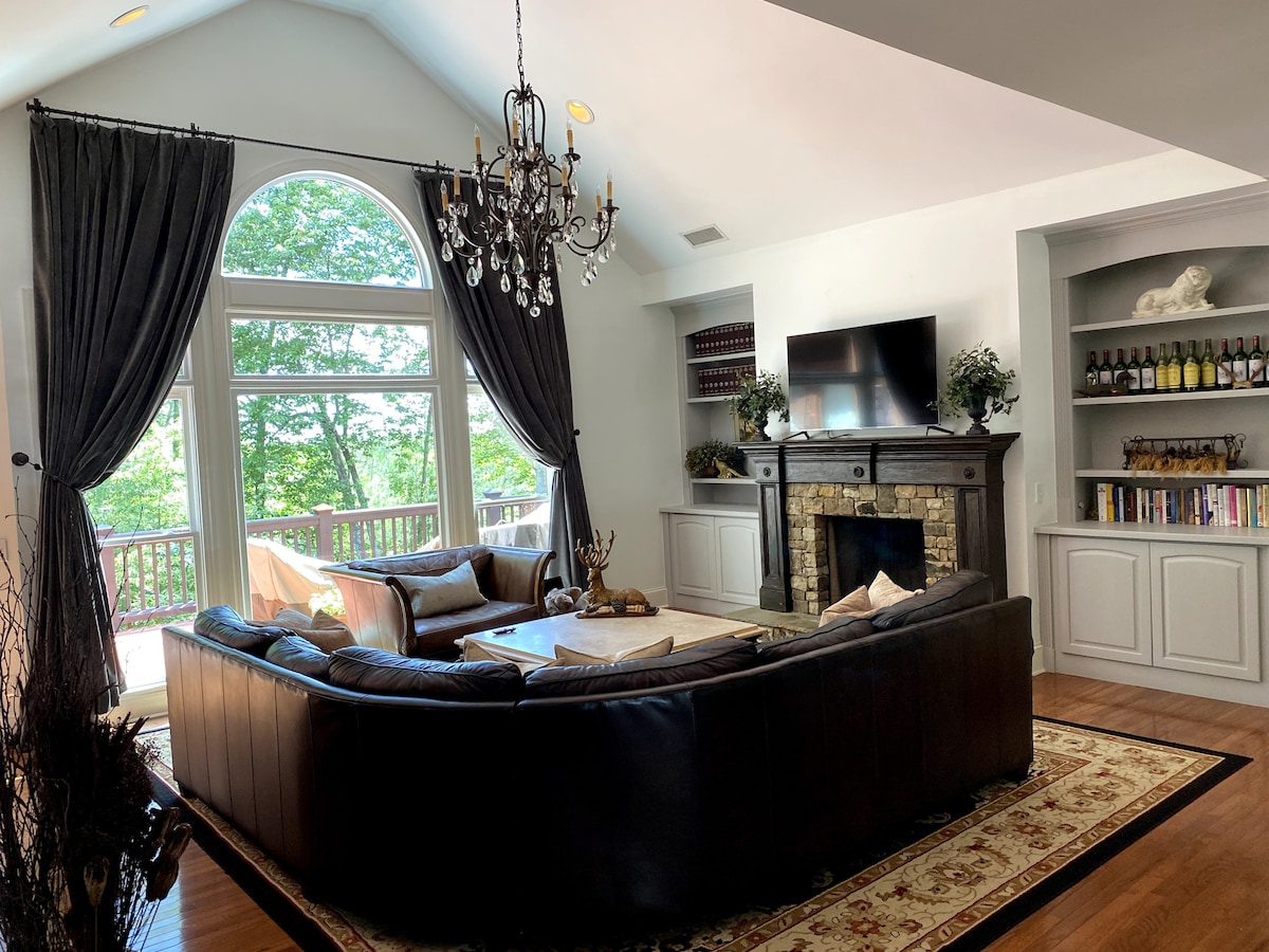 A spacious living room features large windows allowing natural light to illuminate the space. Elegant dark drapes frame the windows, while a chandelier hangs above. Comfortable seating is arranged around a coffee table, complemented by built-in shelves displaying various decorative items and wine bottles.