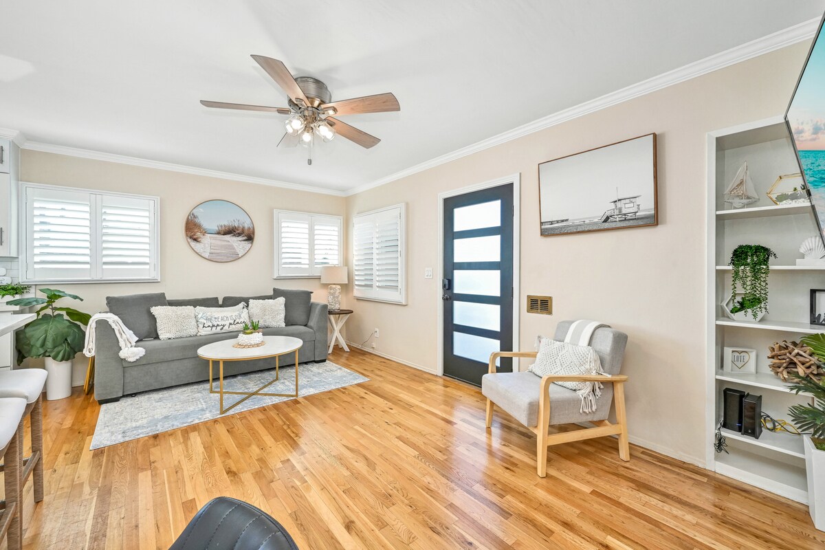 A cozy living area features a gray sofa, a round coffee table, and two armchairs, all arranged on a light rug over hardwood floors. Natural light filters through the windows adorned with shutters, illuminating the space and highlighting decorative wall art.