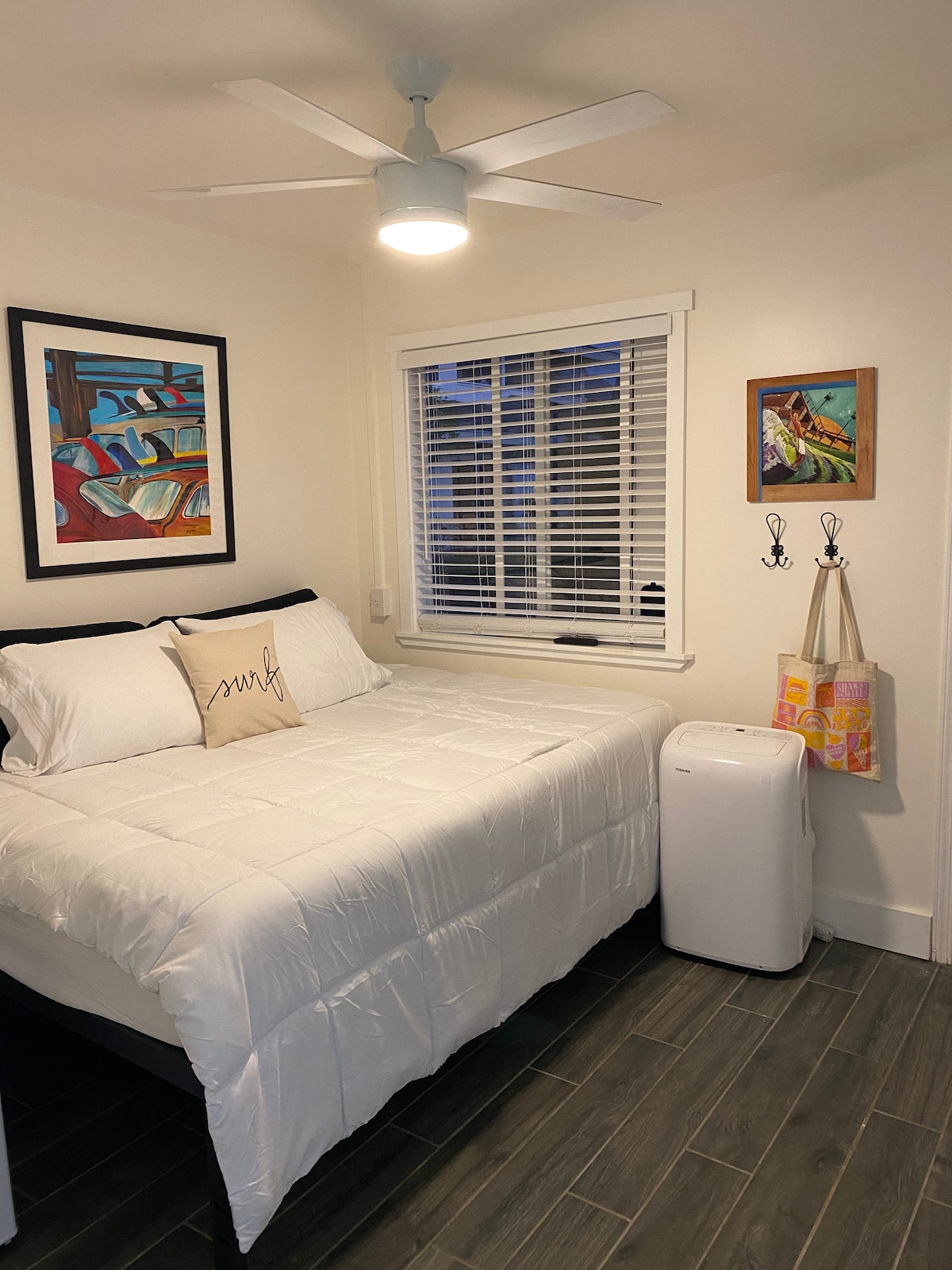 A compact sleeping area features a queen bed topped with a white quilt and decorative pillow. A ceiling fan provides airflow, while a window with blinds allows natural light. Artwork adorns the walls, and a mini-fridge is positioned nearby.
