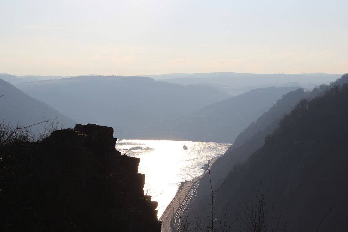 A scenic view of the Rhine River meanders between steep hills, reflecting sunlight on its surface. A small boat can be seen navigating the waterway, with the surrounding landscape showcasing layers of hills and valleys against a soft sky.