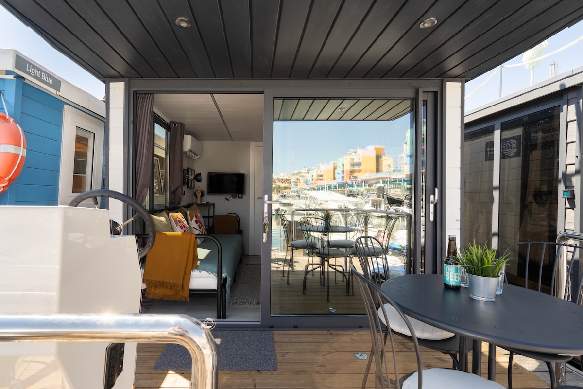 An open living area is visible through large glass doors, revealing a modern interior with a comfortable seating area, television, and decor. An outdoor deck features a table and chairs, with views of the marina and surrounding boats, enhancing the connection to the water.