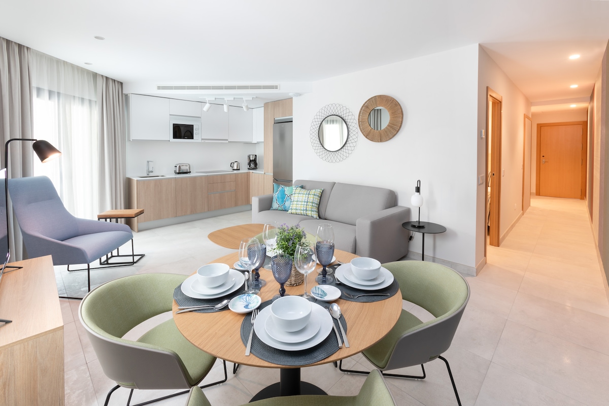 A bright, open living space features a modern dining table set for four, complemented by stylish green chairs. A cozy seating area includes a light grey sofa and a soft blue armchair. Natural light filters through large windows, illuminating the elegant decor.