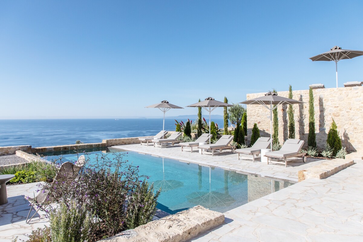 The outdoor area features a shared swimming pool surrounded by sun loungers and umbrellas. Lush greenery and stone walls provide a serene backdrop, while views of the sea enhance the tranquil setting. The pool's water reflects the clear blue sky, inviting relaxation.