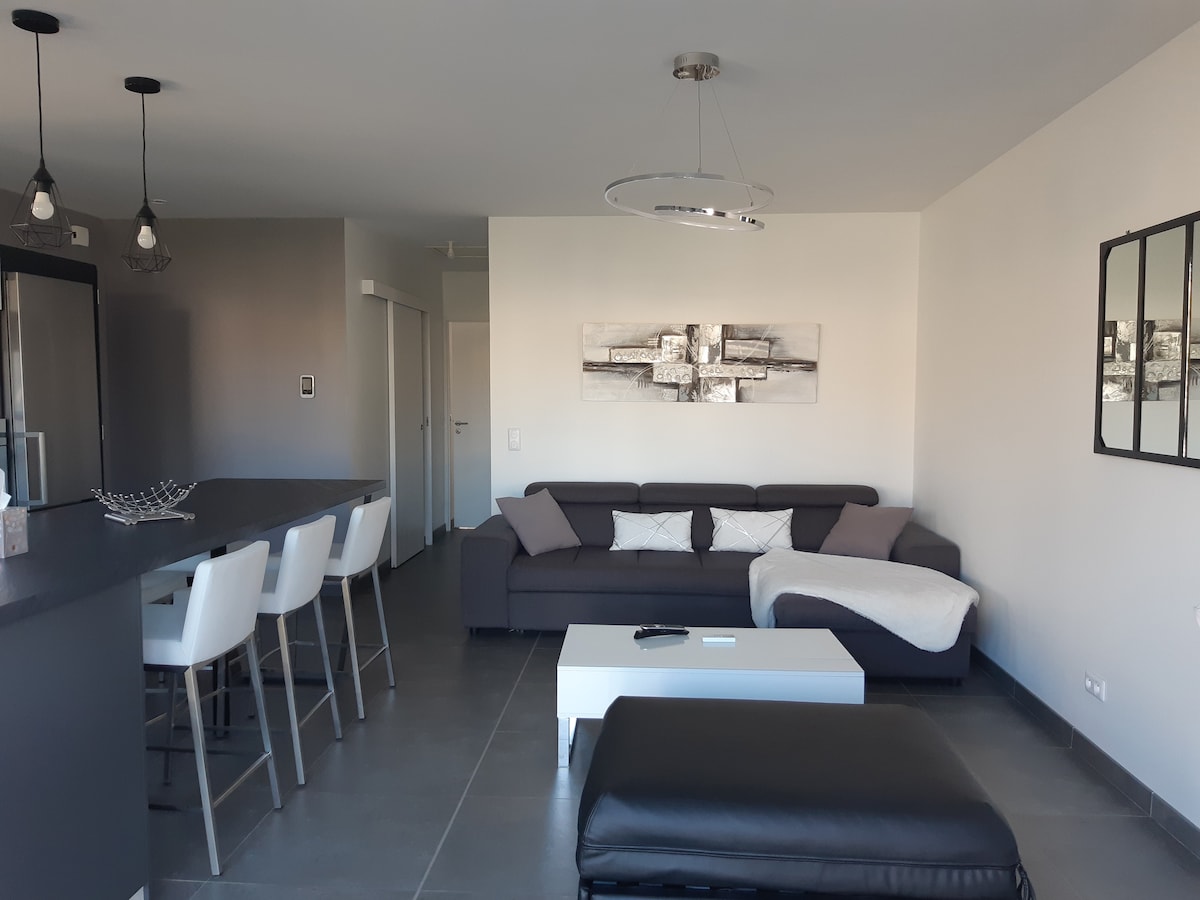 A modern living space is presented, featuring a dark sofa with decorative cushions and a light coffee table at its center. A dining area with high chairs is visible, along with a sleek kitchen. Light fixtures provide a soft ambiance across the open layout.