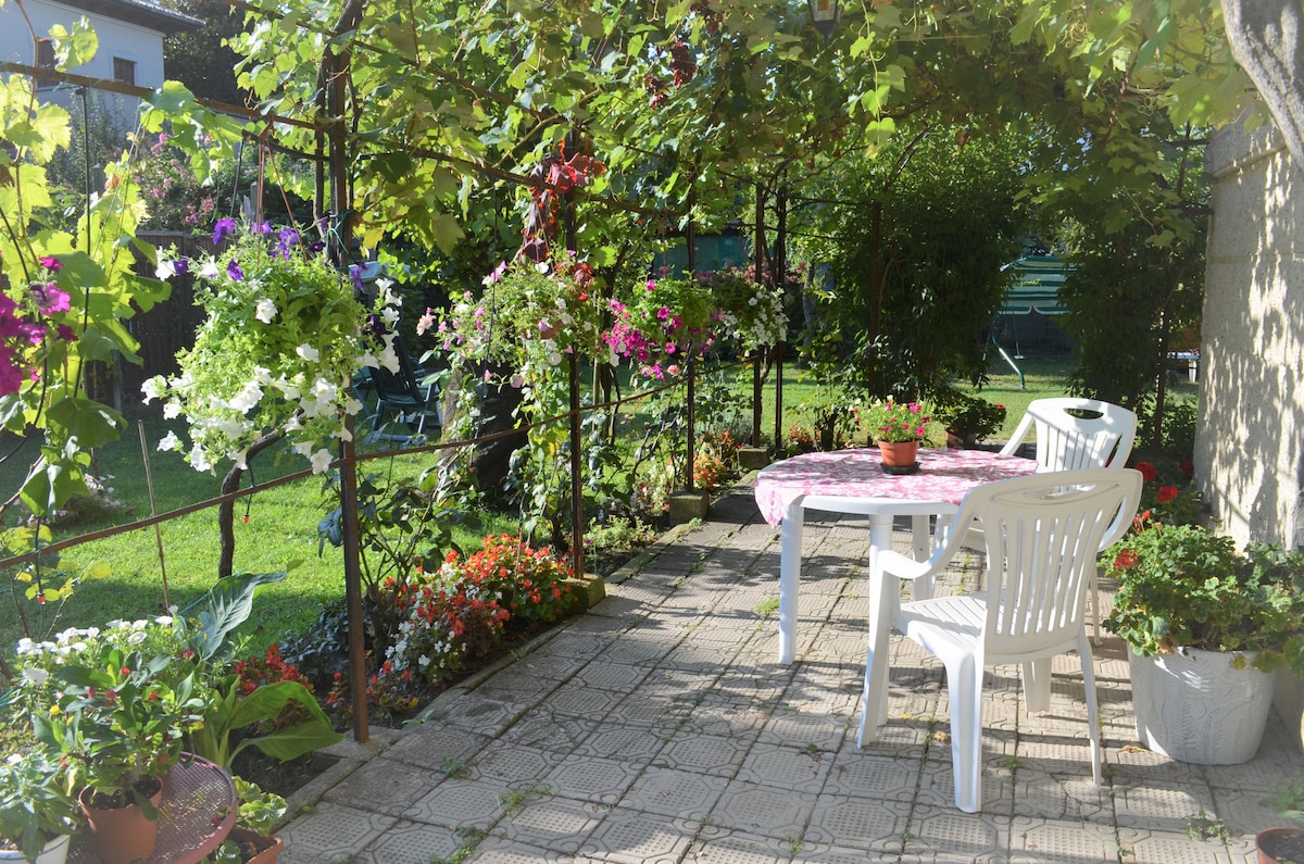 An outdoor patio area is shown, featuring a round table with a pink tablecloth and two white chairs. Surrounding the space are vibrant flowering plants and greenery, creating a serene environment. A well-maintained garden is visible in the background.