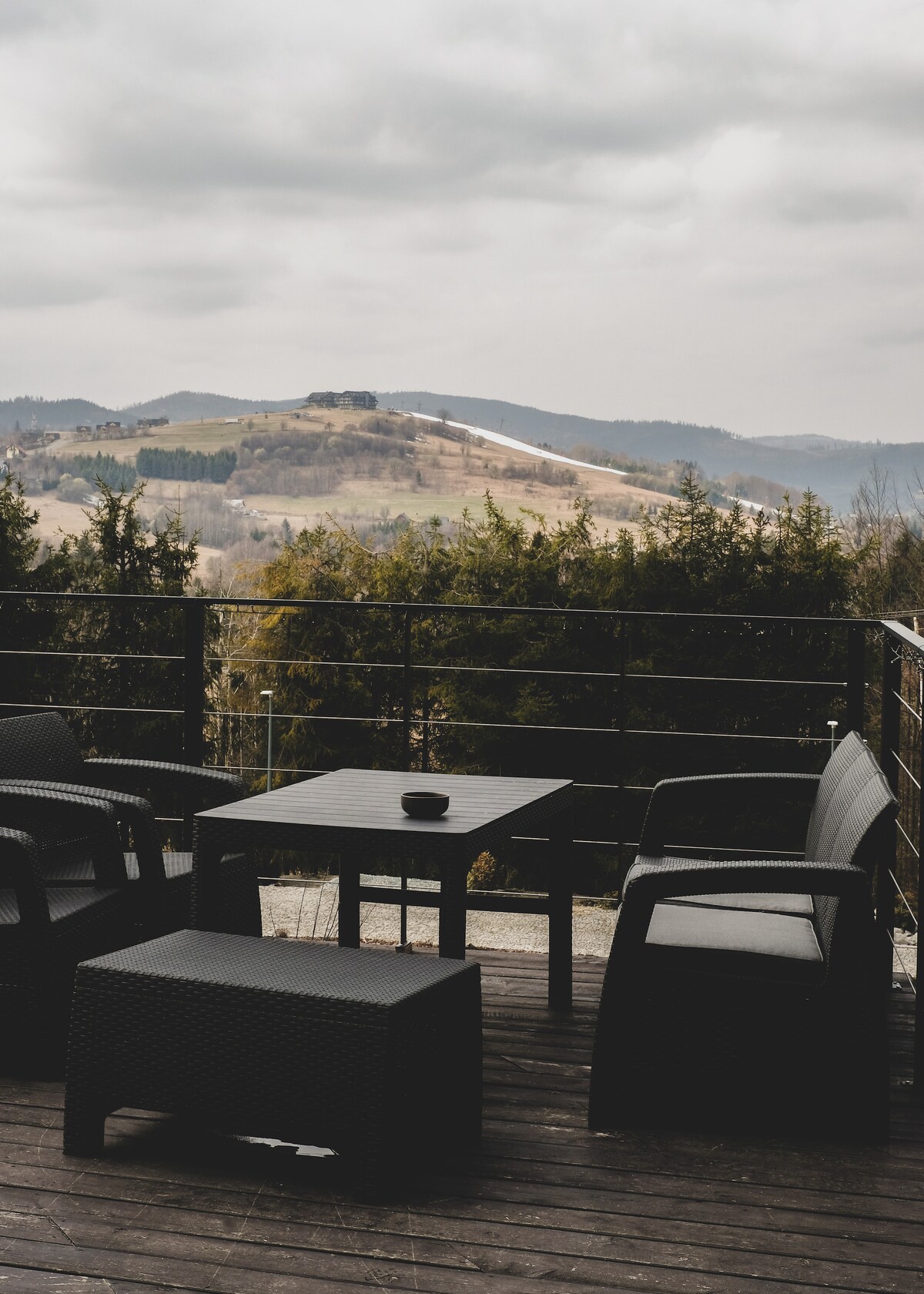A terrace area is furnished with a dark-colored table and chairs, providing a comfortable setting for relaxation. The view features rolling hills in the background, with a mix of greenery and open terrain under a cloudy sky.