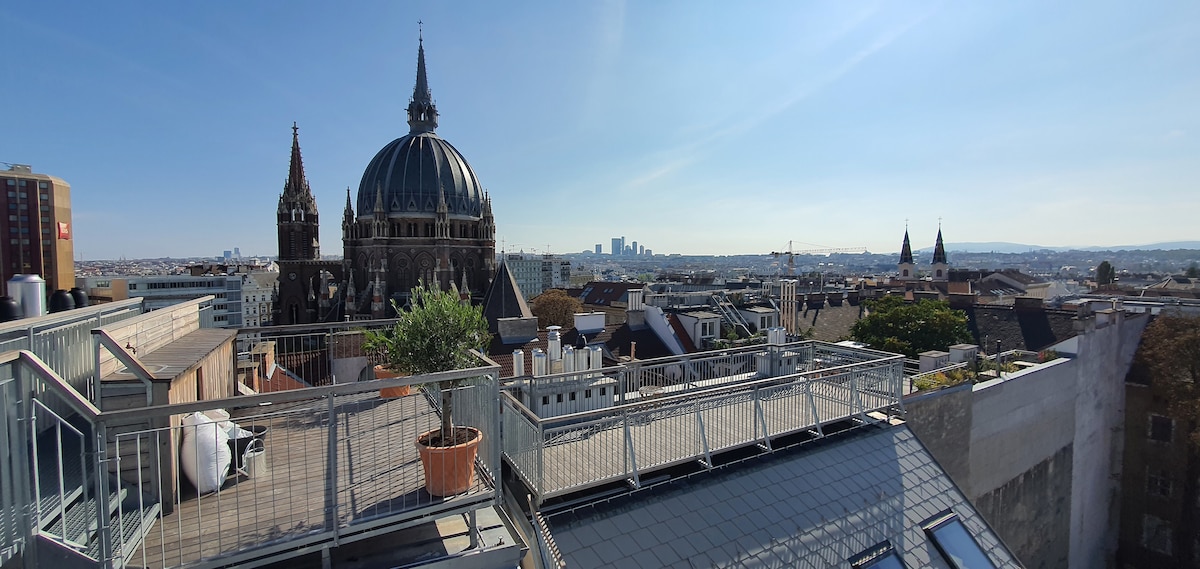 A spacious rooftop terrace offers a panoramic view of the city, featuring notable architectural elements such as domed structures and distant skyline silhouettes. A potted plant is positioned on the terrace, complemented by an extensive railing, providing safety and aesthetic appeal.
