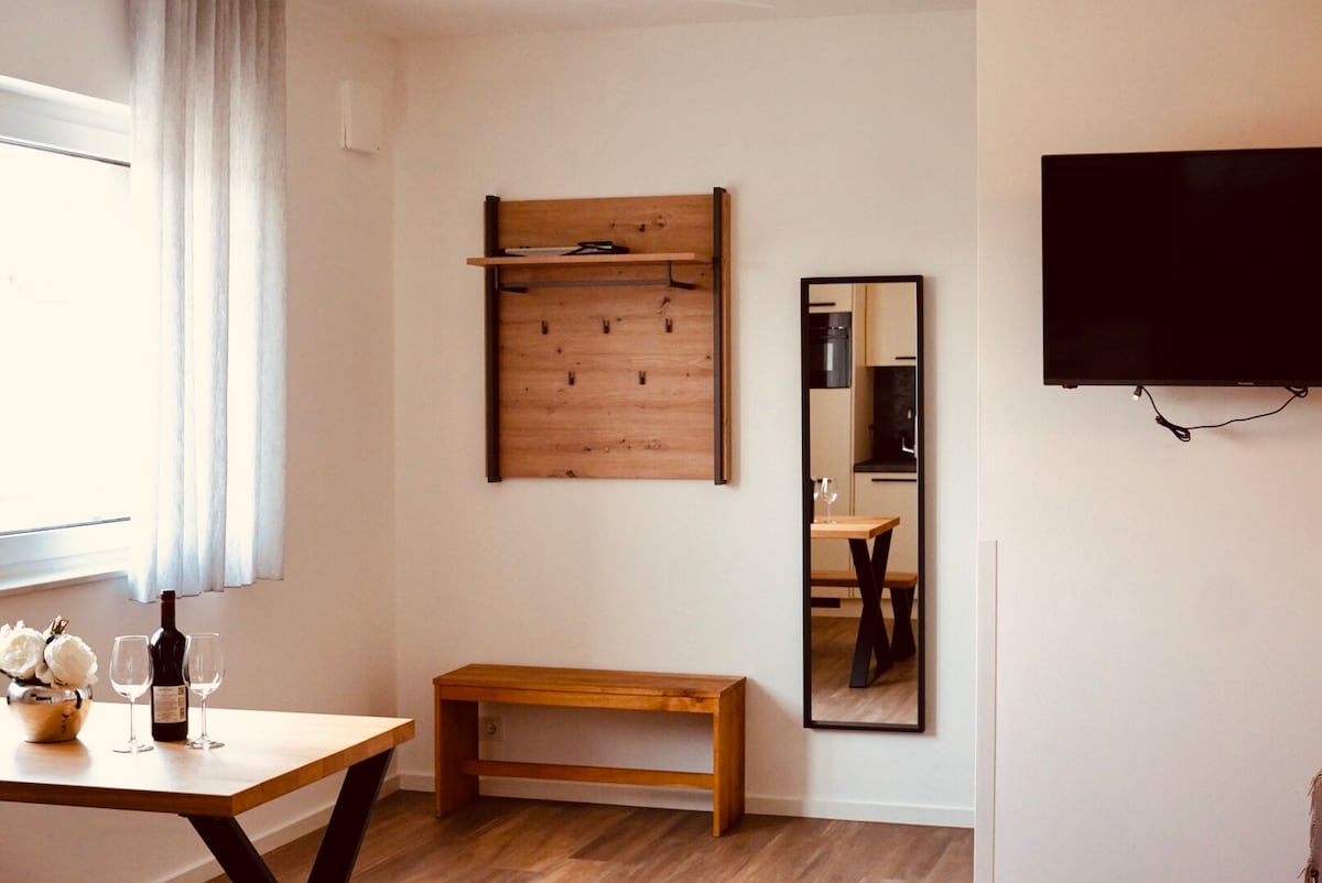 A well-lit area features a wooden table with two wine glasses and a bottle, accompanied by a wooden bench. A full-length mirror is mounted on the wall, and a television is positioned above a shelf. Soft curtains frame the window, enhancing the openness of the space.