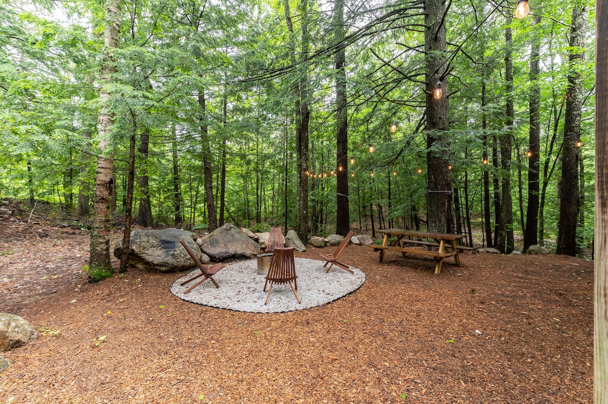 An outdoor seating area is surrounded by a lush forest, featuring a circular stone fire pit with chairs arranged nearby. A rustic picnic table is positioned under the trees, and string lights are draped overhead, creating a cozy ambiance for gatherings.