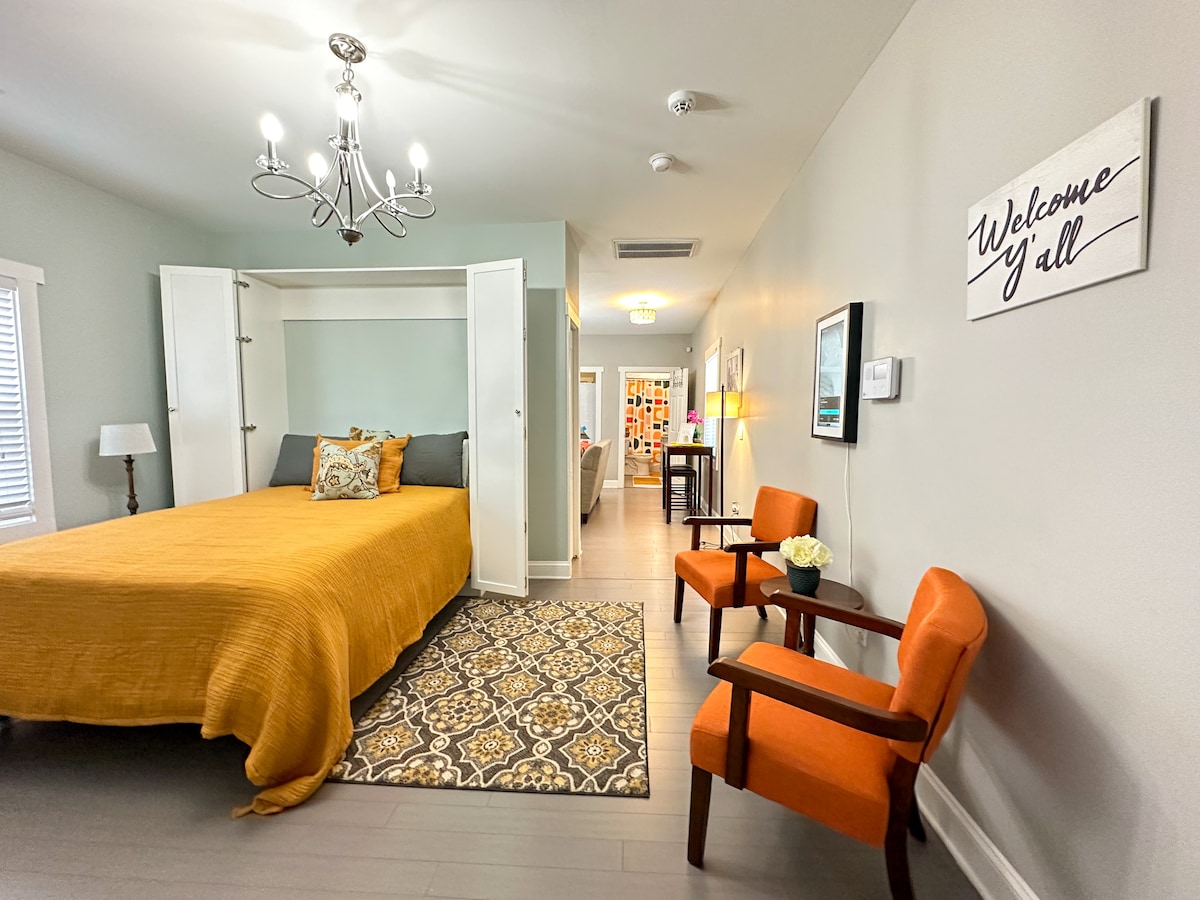A spacious studio features a warm accent wall with an inviting foldable bed covered in a golden-yellow throw. Two orange chairs are positioned by a decorative area rug. Light streams in from a window, illuminating a welcoming sign on the adjacent wall.