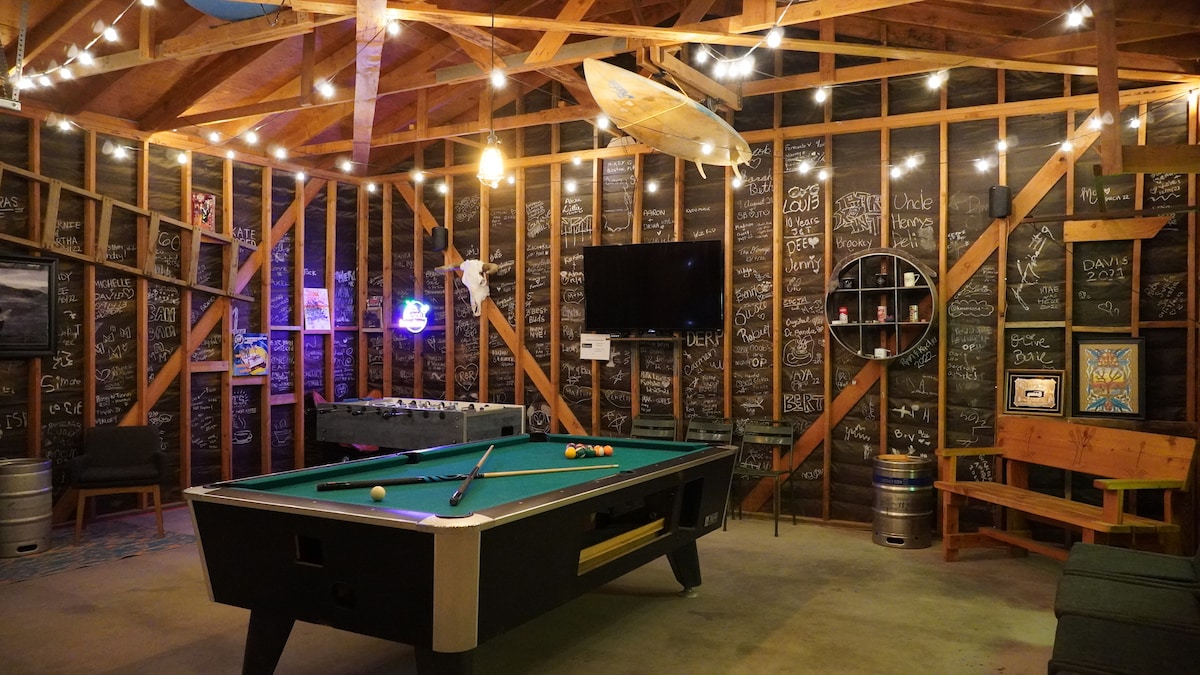 A spacious game room features a green pool table at the center, surrounded by wooden beams and ambient string lights. Decorated with a collection of chalkboard walls displaying various drawings, a 4K TV and additional games are visible, enhancing the entertainment setting.