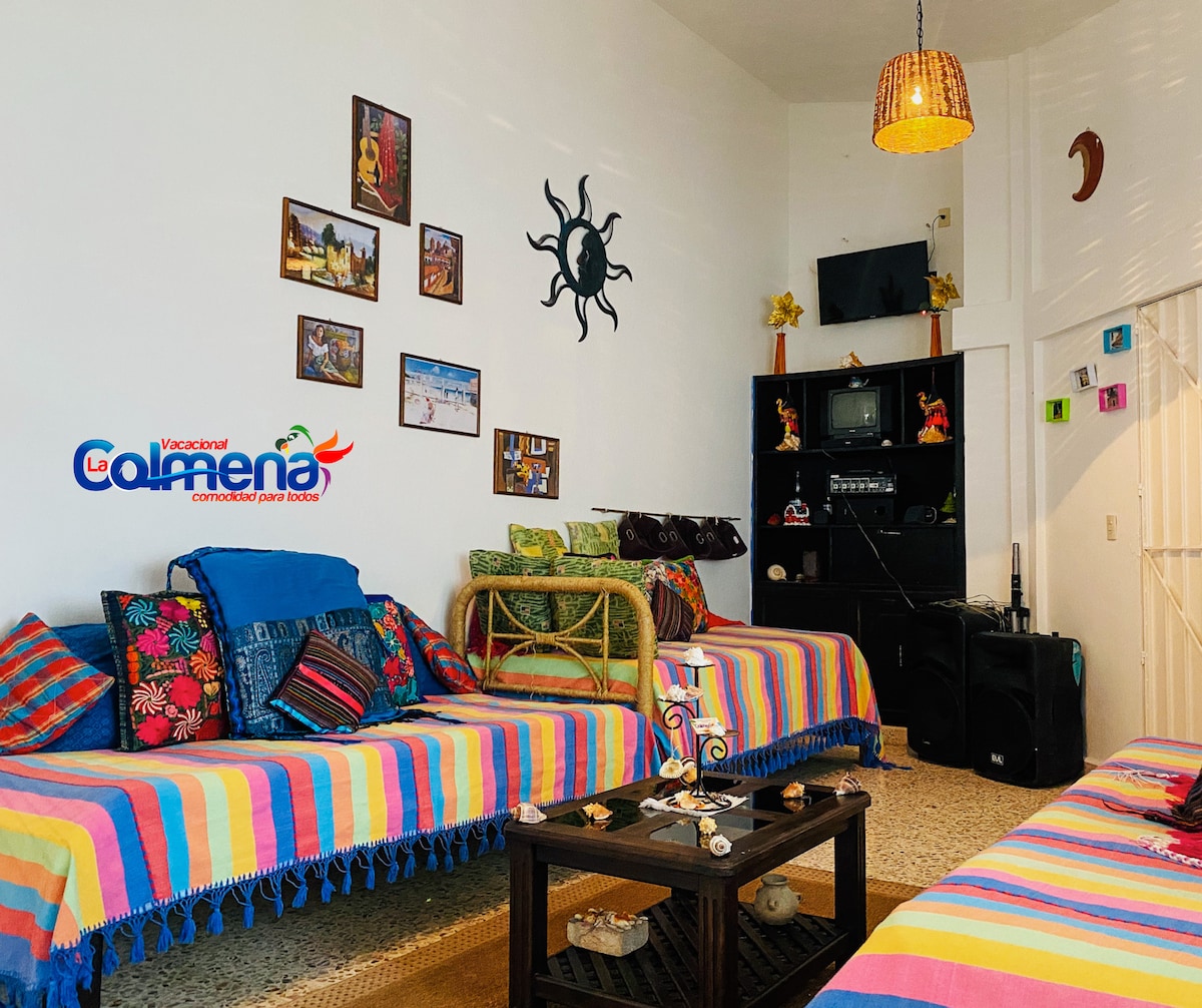A vibrant living area features colorful striped seating with decorative cushions. The walls are adorned with a selection of artwork and photographs, while a dark wood cabinet and a television anchor the space. Natural light fills the room from an adjacent doorway.