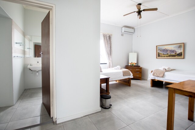 A bright room features two spacious beds positioned against light-colored walls. A wooden table stands next to one bed, and a mirror is visible on a dresser in the corner. Natural light filters in through a window, and a washroom area is seen in the background.