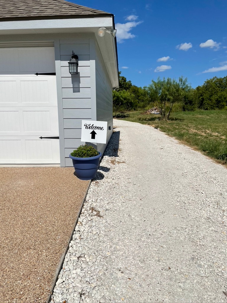 A gravel driveway leads towards a light-colored barn. The corner of the building is visible, along with a welcoming sign placed in a blue pot. Green grass and trees are seen in the background under clear blue skies.