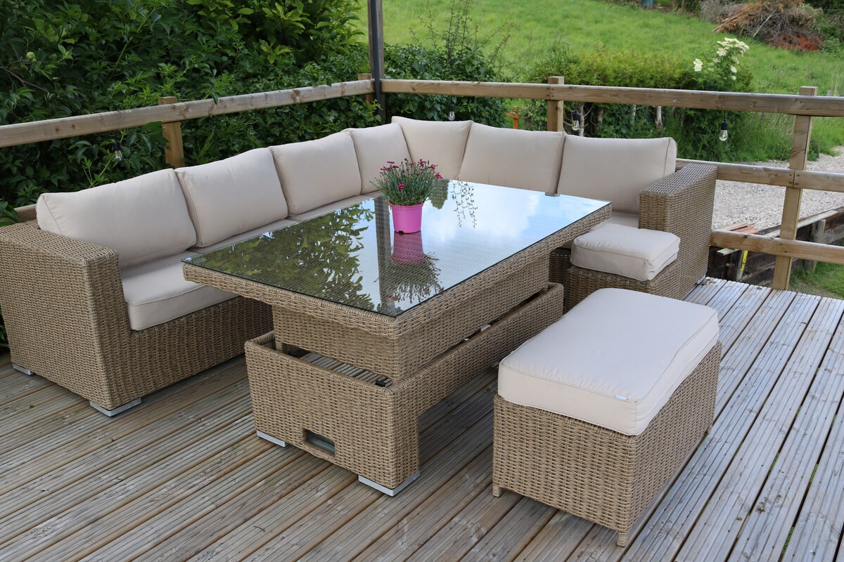 A spacious outdoor seating area features a comfortable sectional sofa with soft beige cushions, arranged around a rectangular glass-top table. Two matching ottomans are positioned nearby, and vibrant greenery lines the background, enhancing the tranquil environment.