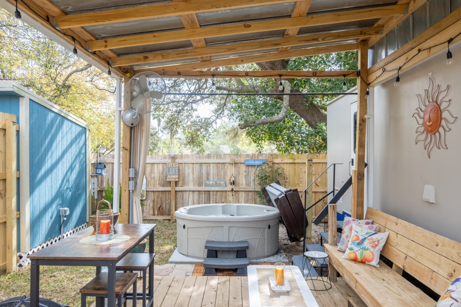 An outdoor covered deck features a hot tub surrounded by wooden fencing. A small table and bench provide seating, with colorful cushions for added comfort. Natural light filters through the space, highlighting the relaxing atmosphere and leafy trees visible in the background.