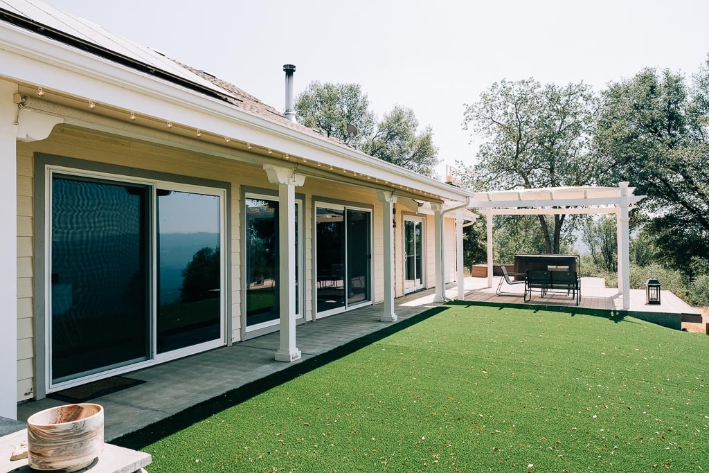 A spacious outdoor area features a green lawn, surrounded by trees. Large glass doors provide access to the indoor living space. An overhead pergola shades a dining area, with a table and chairs set up for outdoor meals.