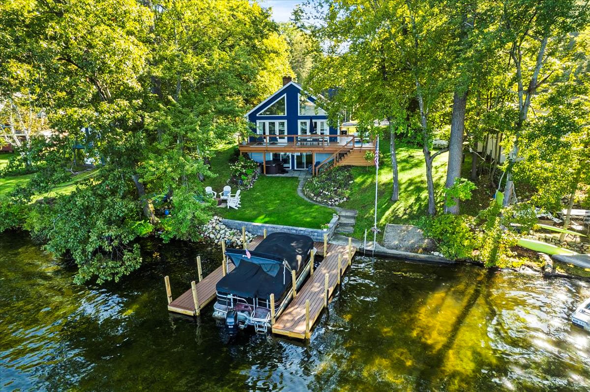 An aerial view captures the home set among lush greenery, featuring a large deck and direct access to the lake. A private dock with a covered boat is visible, while spacious lawns provide room for outdoor activities. The inviting facade boasts multiple large windows.