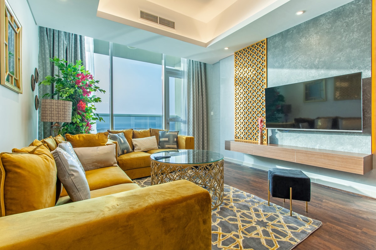A modern living space is showcased with a plush yellow sofa and complementary pillows. A sleek glass coffee table rests on a patterned rug, while a large television is mounted on a textured wall. Natural light filters through expansive windows adorned with elegant curtains.