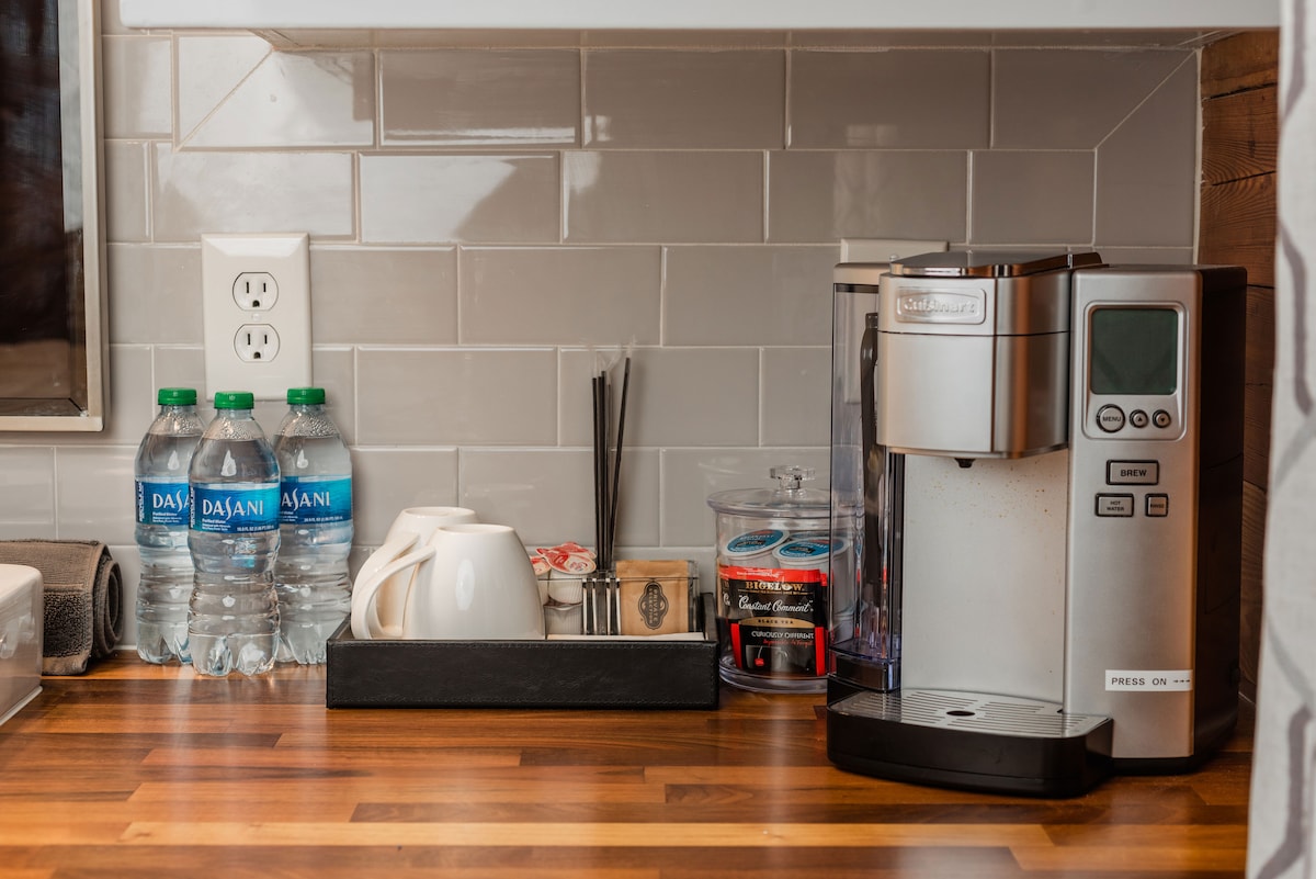 A compact kitchen counter displays a Keurig coffee maker beside bottled water and various coffee accessories. A set of neatly arranged cups and a container filled with tea bags are visible, alongside decorative items such as straws. The backsplash features modern gray tiles.