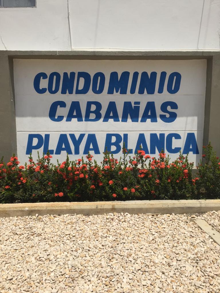 A prominent sign displaying the name 'Condominio Cabañas Playa Blanca' is set against a white wall. Bright red flowers border the sign, contrasting with the gravel pathway that leads up to it. The overall setting is part of a well-maintained area.