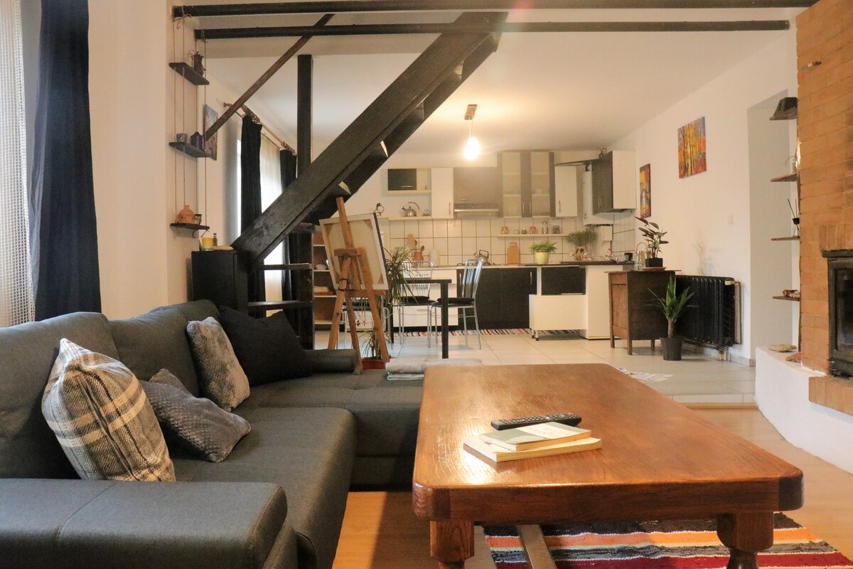 A spacious living area features a dark, L-shaped sofa adorned with patterned cushions, complemented by a large wooden coffee table. The open kitchen is visible in the background, showcasing modern appliances and a dining area. Light enters through large windows, illuminating the room.