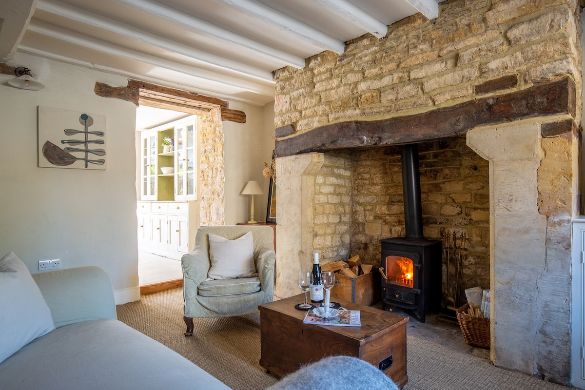 A cozy sitting room features a stone inglenook fireplace with a wood burning stove, providing warmth. A comfortable armchair sits next to a wooden coffee table, while exposed Cotswold stone walls and oak beams highlight the cottage's original character.