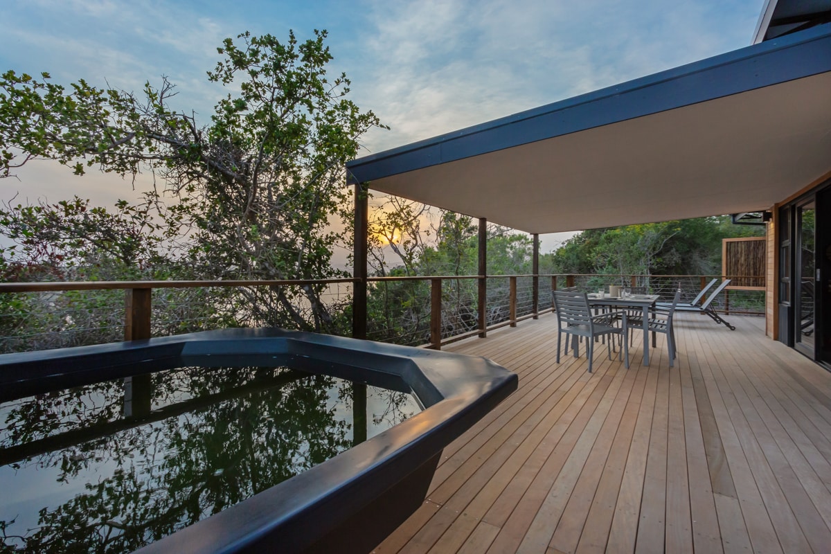 An expansive patio features a wood-fired hot tub and a dining area with a table and chairs. Sun-loungers are positioned nearby, surrounded by greenery. The scene is softened by the warm glow of sunset in the background.