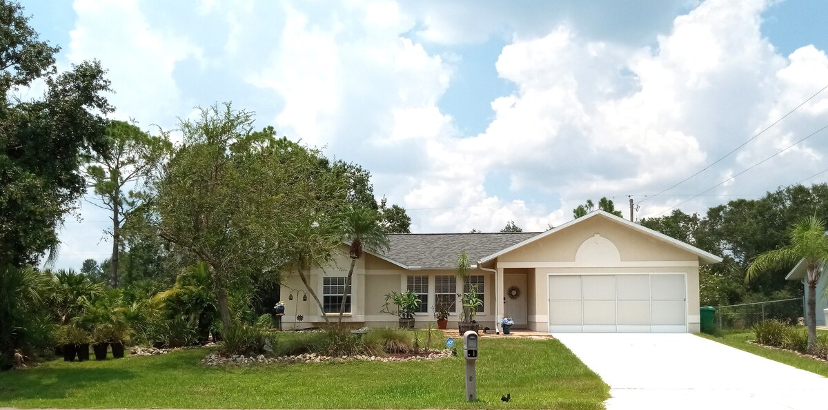 A single-story home is displayed, featuring a well-maintained garden with various plants. The driveway leads to a spacious garage. Sunlight illuminates the exterior, highlighting large windows that provide views into the living spaces. A peaceful natural landscape surrounds the property.
