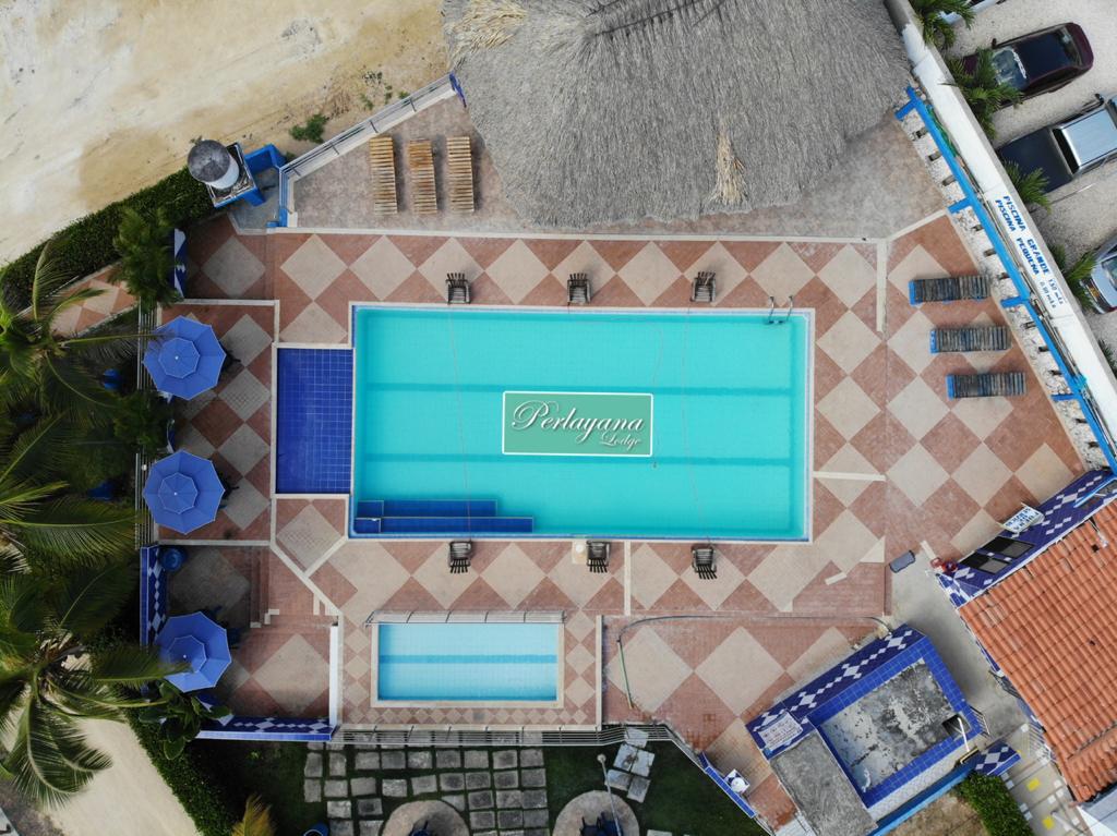 An aerial view captures a large swimming pool surrounded by tiled flooring and sun loungers. A smaller jacuzzi is visible nearby. Several umbrellas provide shade, and a thatched-roof structure adds to the outdoor space's charm. The vibrant blue of the pool complements the surrounding area.