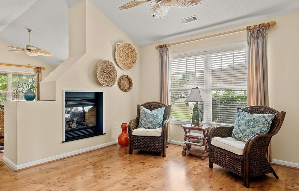A bright living space features two wicker chairs adorned with decorative pillows, positioned beside a lamp on a small table. Natural light floods through a window with sheer curtains, and a fireplace is built into the wall, accompanied by woven wall art.