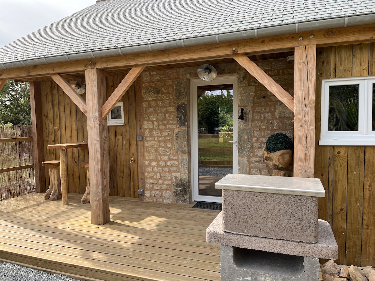 The entrance to a wooden cottage is framed by a rustic porch. Stone and wood accents create a charming exterior. A small table with two stools and a decorative planter are located nearby, inviting relaxation. Natural light highlights the entrance door, which is complemented by a sleek window.