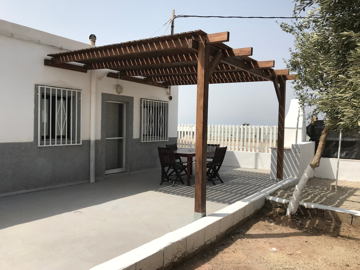 An outdoor patio area features a wooden pergola providing shade over a table with four matching chairs. The ground is paved, and a modest garden area is visible alongside the structure, which includes a white wall and a fence in the background.